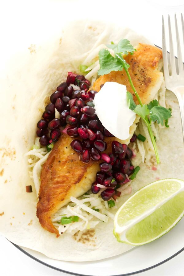 Fish taco with slaw, pomegranate seeds, cilantro, sour cream, and lime wedges.