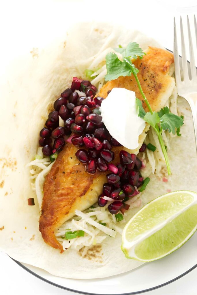 Fish taco with slaw, pomegranate seeds, cilantro, sour cream, and lime wedges.