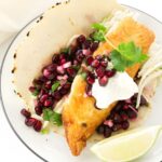Fish taco with pomegranate, herbs, cabbage, white sauce, and lime wedge.