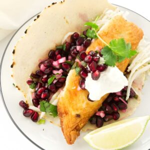 Fish taco with pomegranate, herbs, cabbage, white sauce, and lime wedge.
