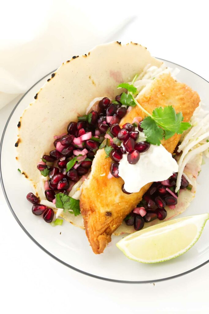 Fish taco with pomegranate, herbs, cabbage, white sauce, and lime wedge.