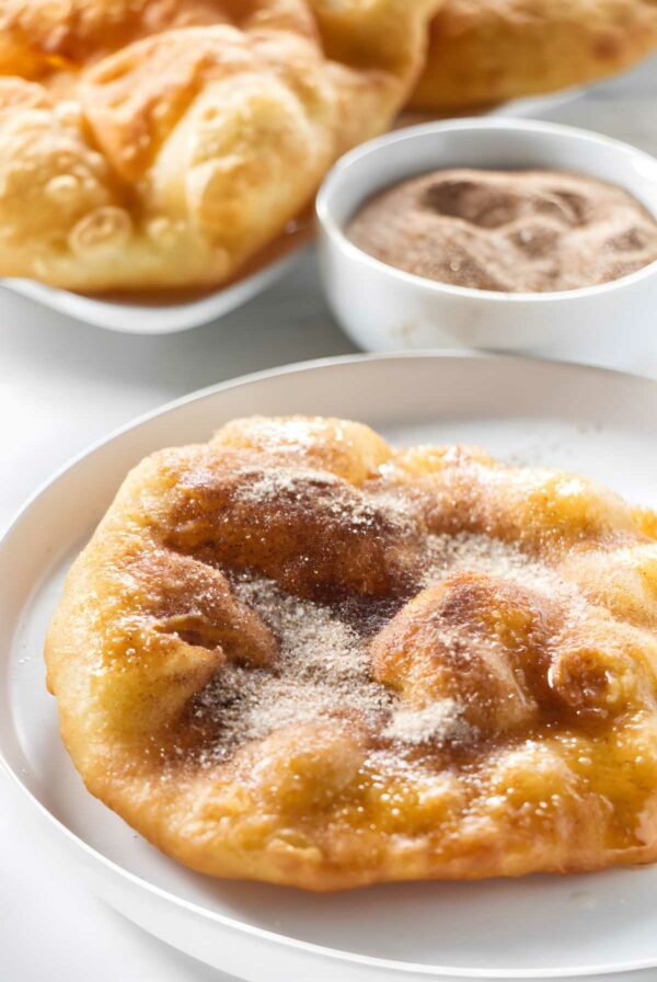 Sugary fried bread on white plate with bowl of cinnamon sugar behind.
