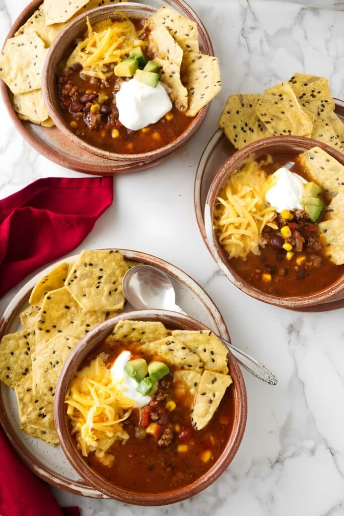 Three chili bowls with toppings, tortilla chips, red napkin, and spoons.
