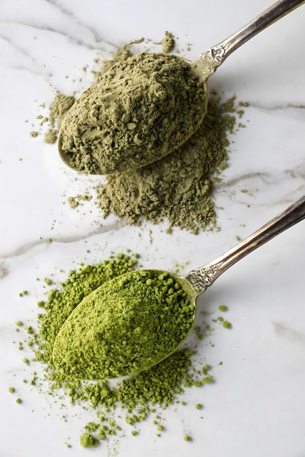 Two metal spoons with matcha powder on white marble surface.