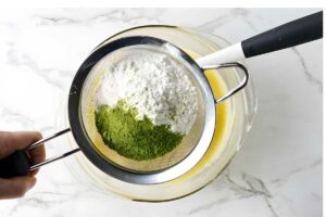 Hand sifting powdered sugar and matcha into yellow mixture in glass bowl.
