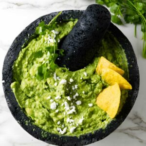 Stone mortar with guacamole, tortilla chips, cheese, limes, and cilantro.