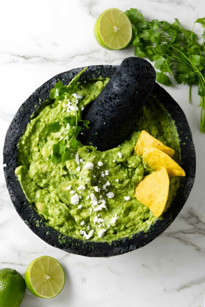 Stone mortar with guacamole, tortilla chips, cheese, limes, and cilantro.