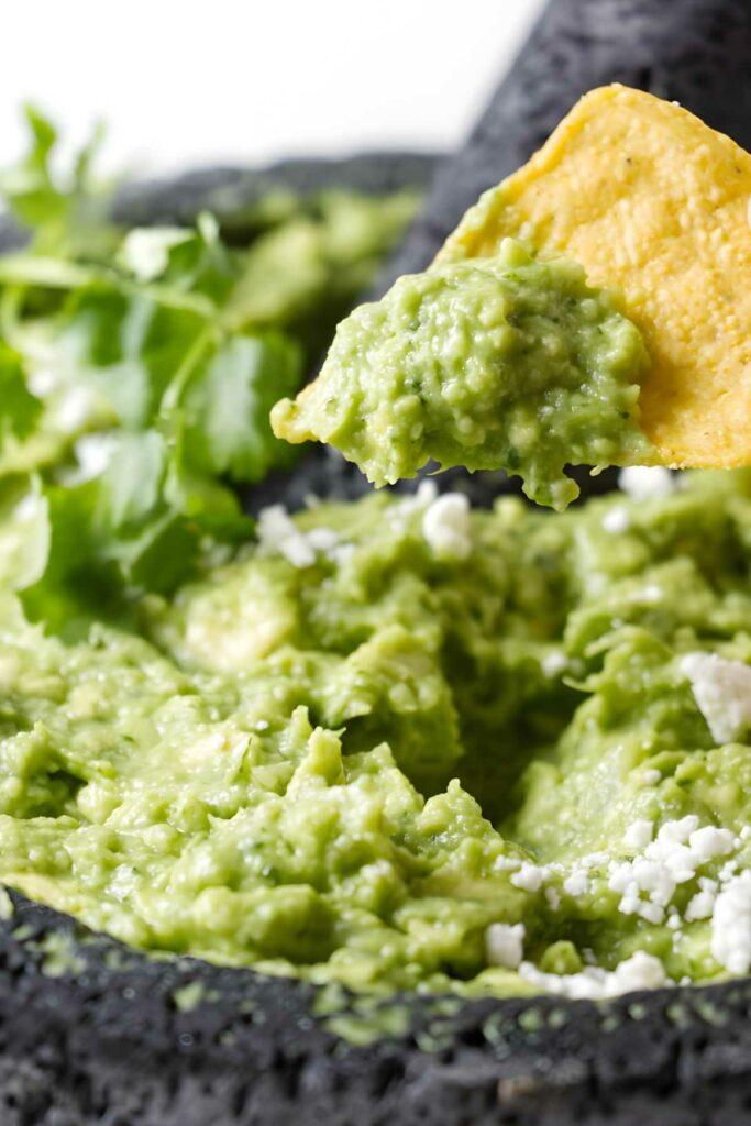 Tortilla chip dipping into guacamole topped with cilantro and crumbled cheese.