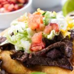 Close-up of fry bread taco with beef, toppings, sliced avocado, and bowls.