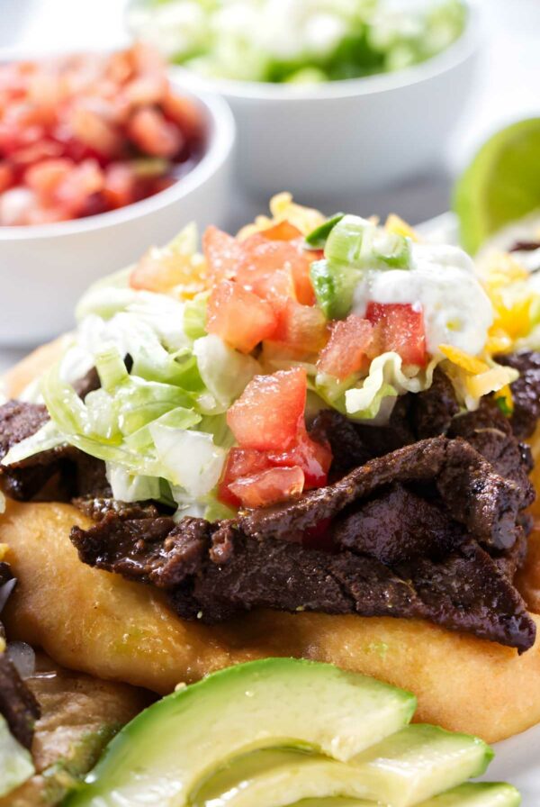 Close-up of fry bread taco with beef, toppings, sliced avocado, and bowls.