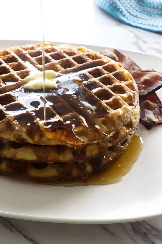 Stack of sourdough waffles with butter, syrup pouring, served with crispy bacon.