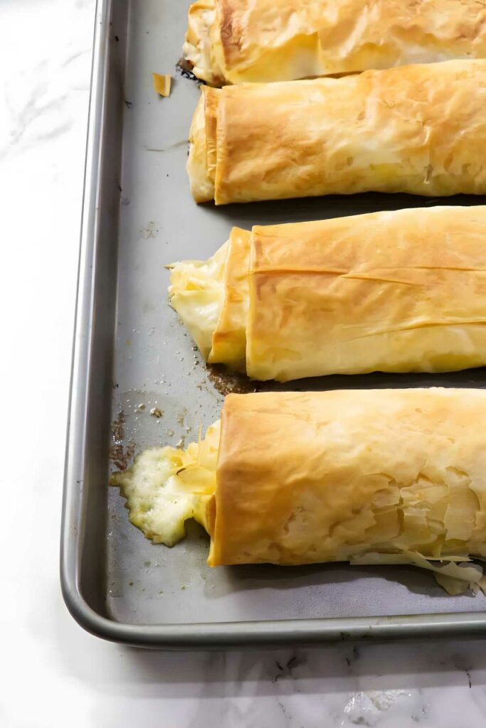Four golden brown cheese-filled phyllo rolls on a baking sheet.