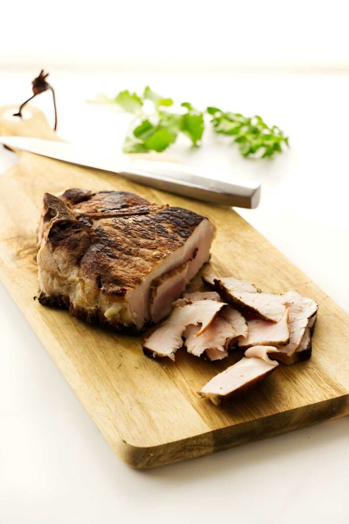 Sliced pork on cutting board with knife and fresh herbs.
