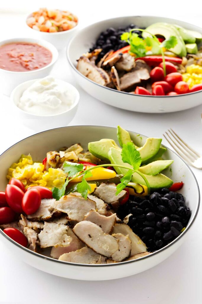 Two tex mex fajita bowls with pork and veggies. Sour cream and salsa in the back. 