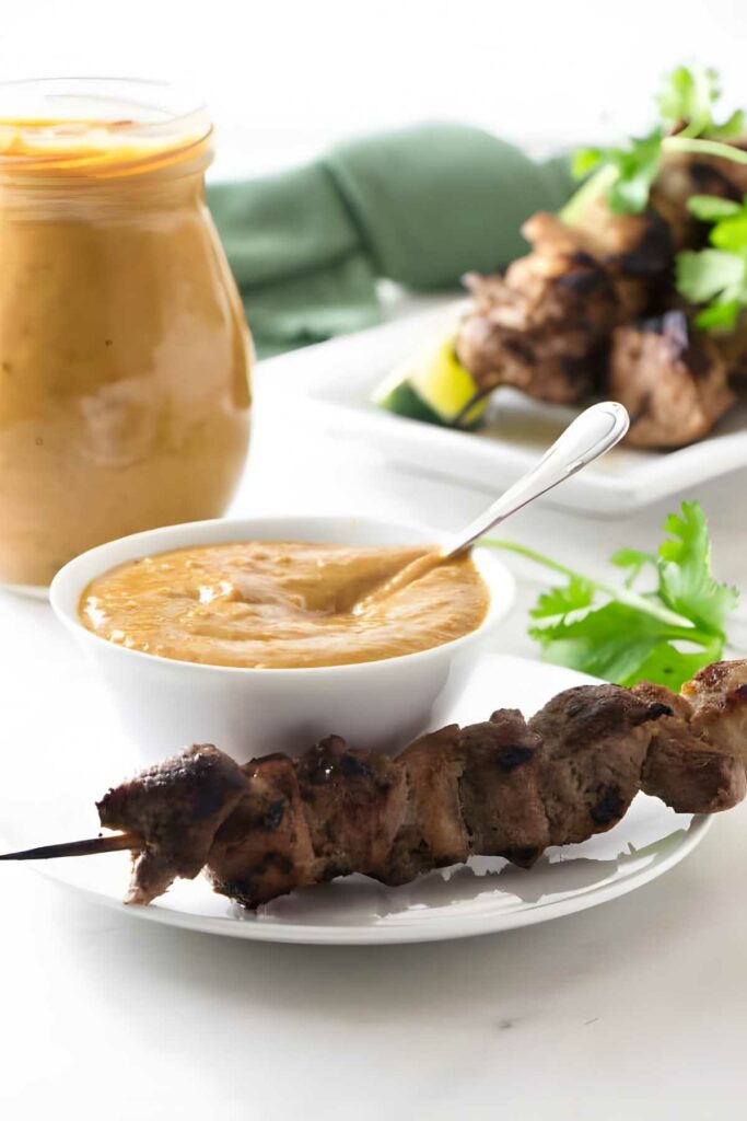 Grilled pork skewers with peanut sauce, fresh herbs, and sauce jar.