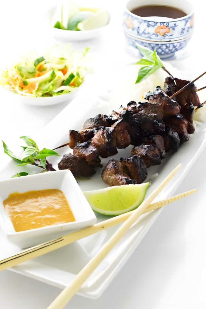 Grilled pork satay skewers with lime, sauce, herbs; salad and tea behind.