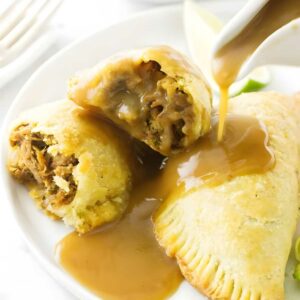 Two golden-brown empanadas with gravy, one cut open, lime slices behind.