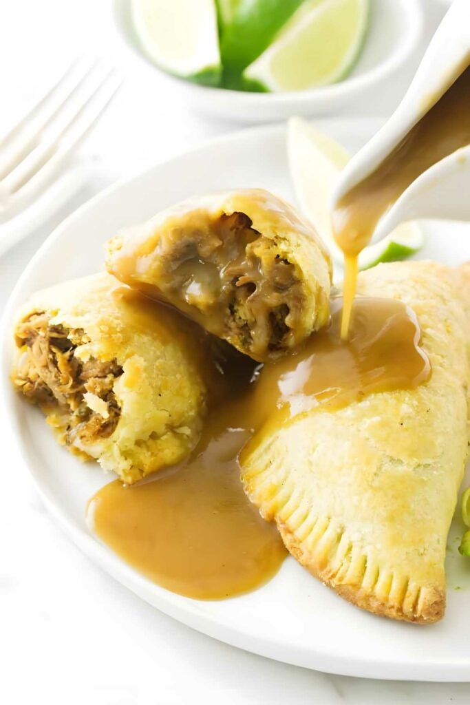 Two golden baked empanadas with gravy, one cut open, lime slices behind.
