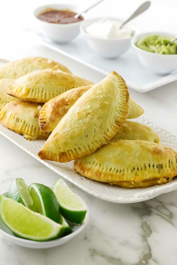 Plate of golden baked empanadas with salsas, avocado, and lime wedges.