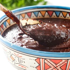 Spoon lifting dark mole sauce from colorful patterned bowl, herbs blurred behind.