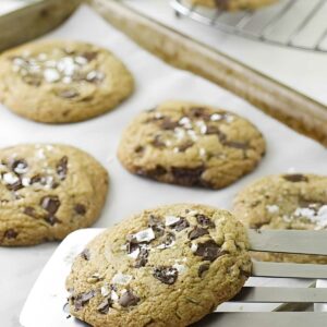 Spatula lifts large chocolate chip cookie with sea salt from baking sheet.