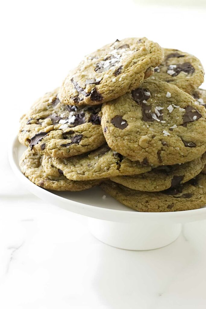 Stack of chocolate chip cookies with sea salt on white cake stand.
