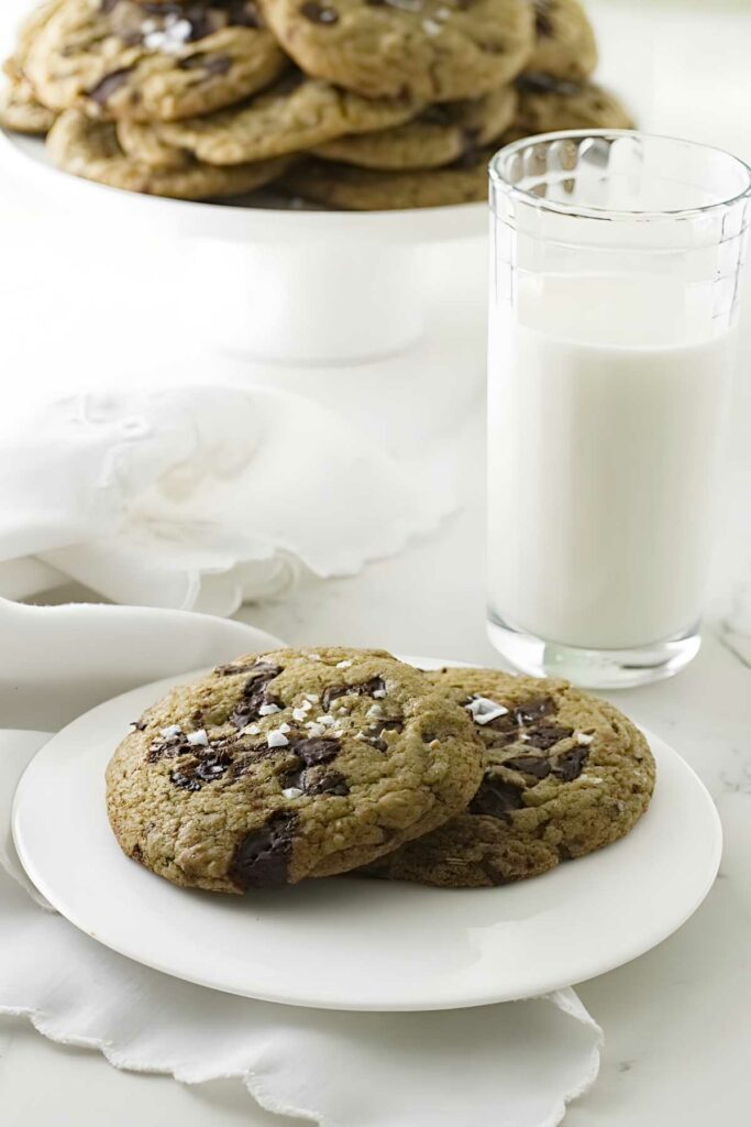 Two sea salt chocolate chip cookies and glass of milk on white plate.