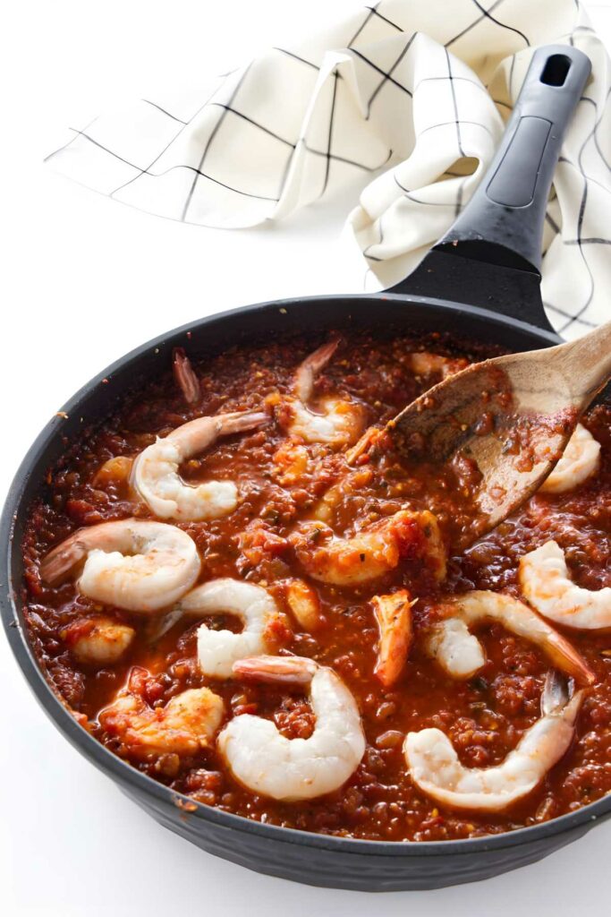 Shrimp cooking in spicy tomato sauce in a skillet, stirred with wooden spoon.