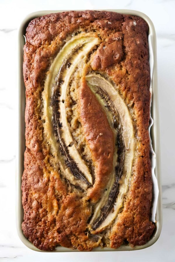 Golden banana bread loaf with sliced bananas on top in a baking pan.