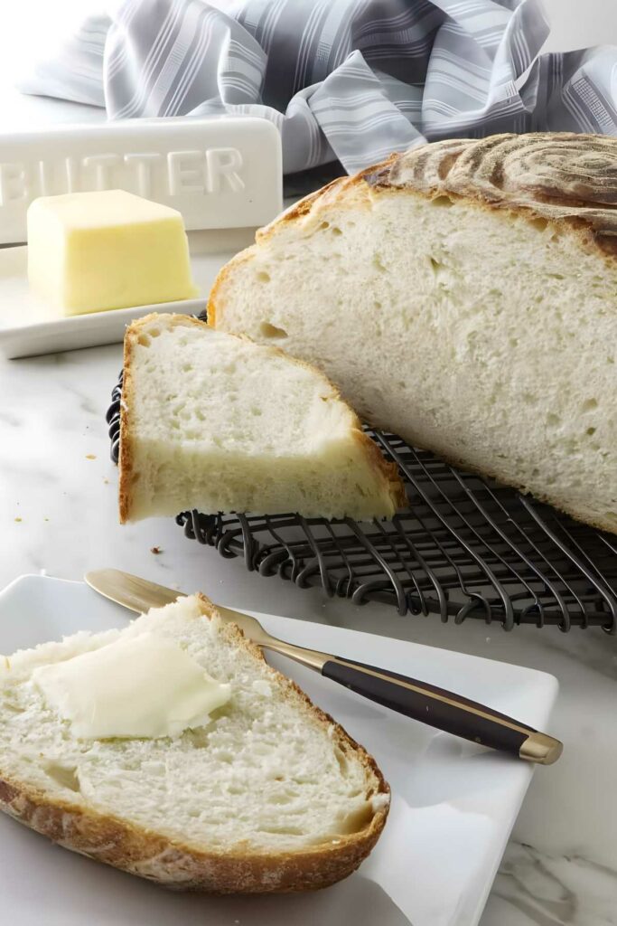 Freshly baked sourdough bread with buttered slice, knife, and butter on counter.