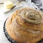 Round rustic sourdough bread loaf on cooling rack, butter and towel in background.