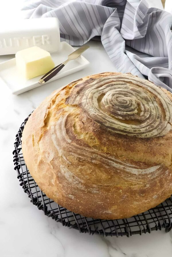Round rustic sourdough bread loaf on cooling rack, butter and towel in background.