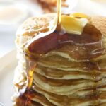 Stack of pancakes with butter and syrup, bacon and eggs in background.