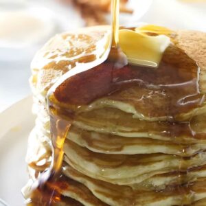 Stack of pancakes with butter and syrup, bacon and eggs in background.