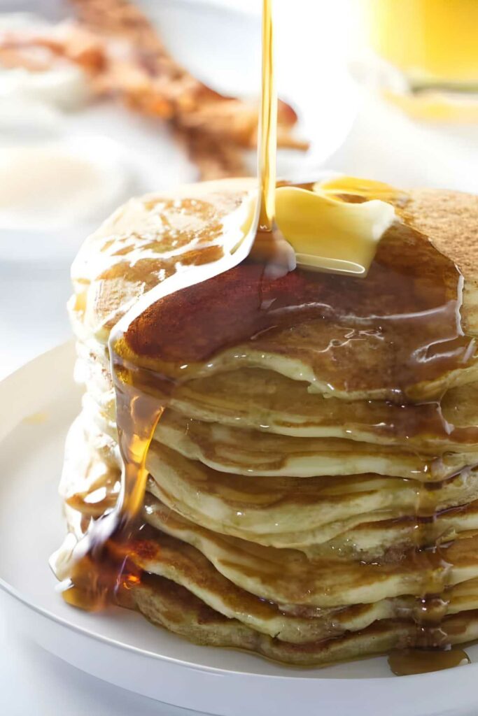 Stack of sourdough pancakes with butter and syrup, bacon and eggs in background.