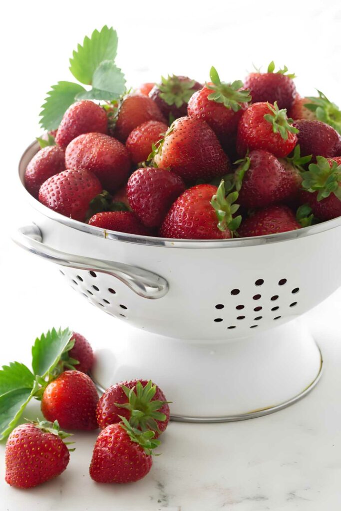 White colander of fresh strawberries with scattered berries.