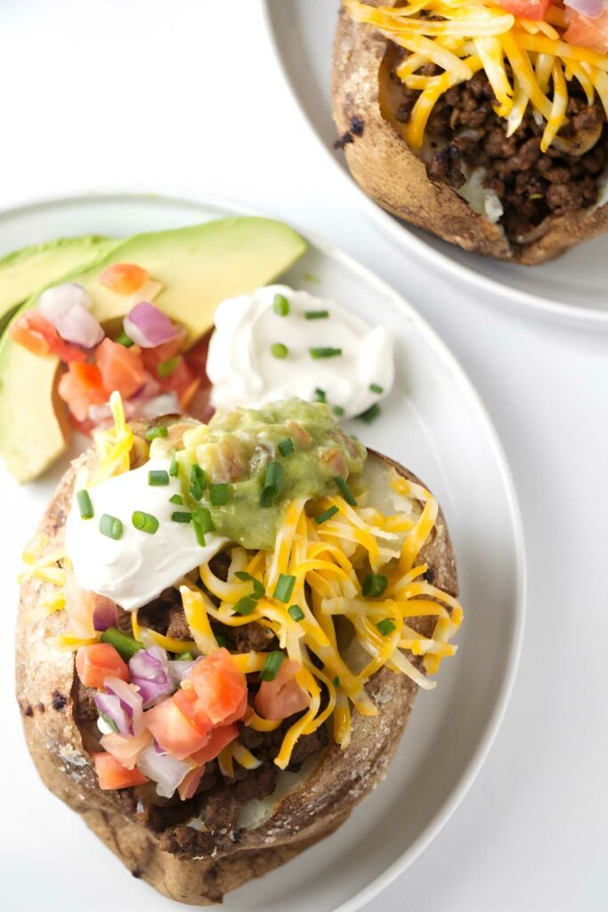 A taco stuffed baked potato on a plate with sour cream and avocado slices.