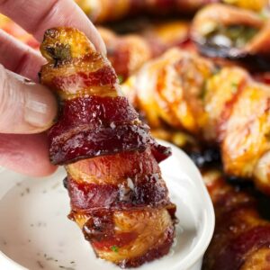Hand dipping bacon-wrapped jalapeño popper into white sauce, others behind.
