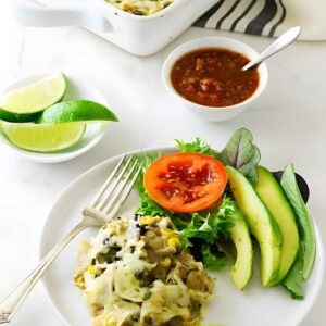 Cheesy casserole plate with avocado, lettuce, tomato, lime, and background salsa.