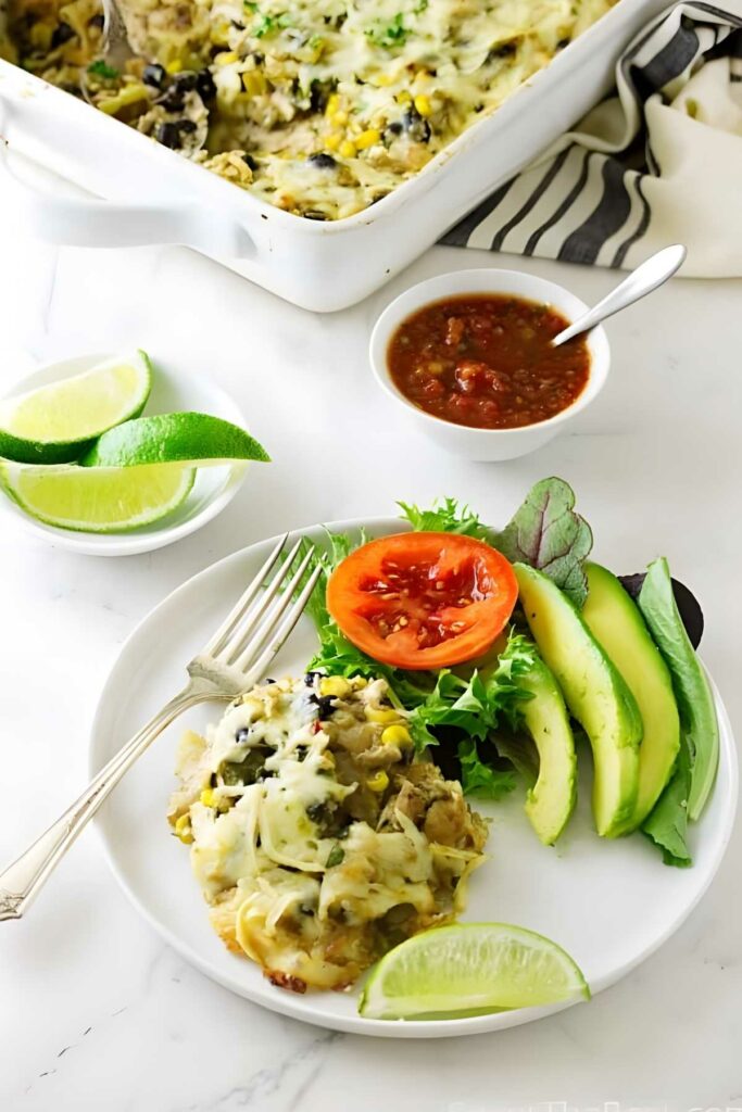 Cheesy casserole plate with avocado, lettuce, tomato, lime, and background salsa.
