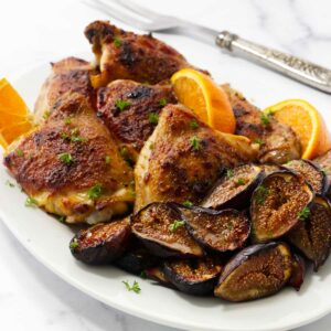Roasted chicken and figs on a serving plate with orange wedges.