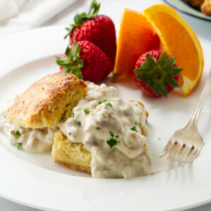 A flaky biscuit covered in sausage gravy on a plate with fruit.