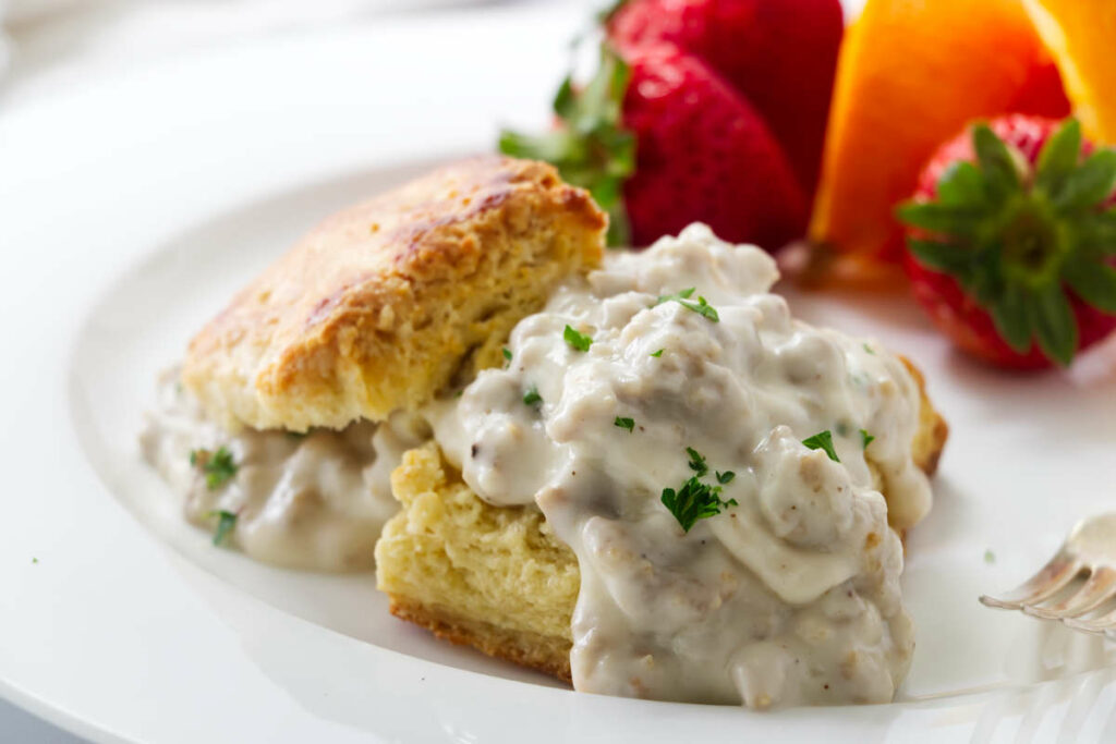 A biscuit with gravy on a plate with a fork and fruit.