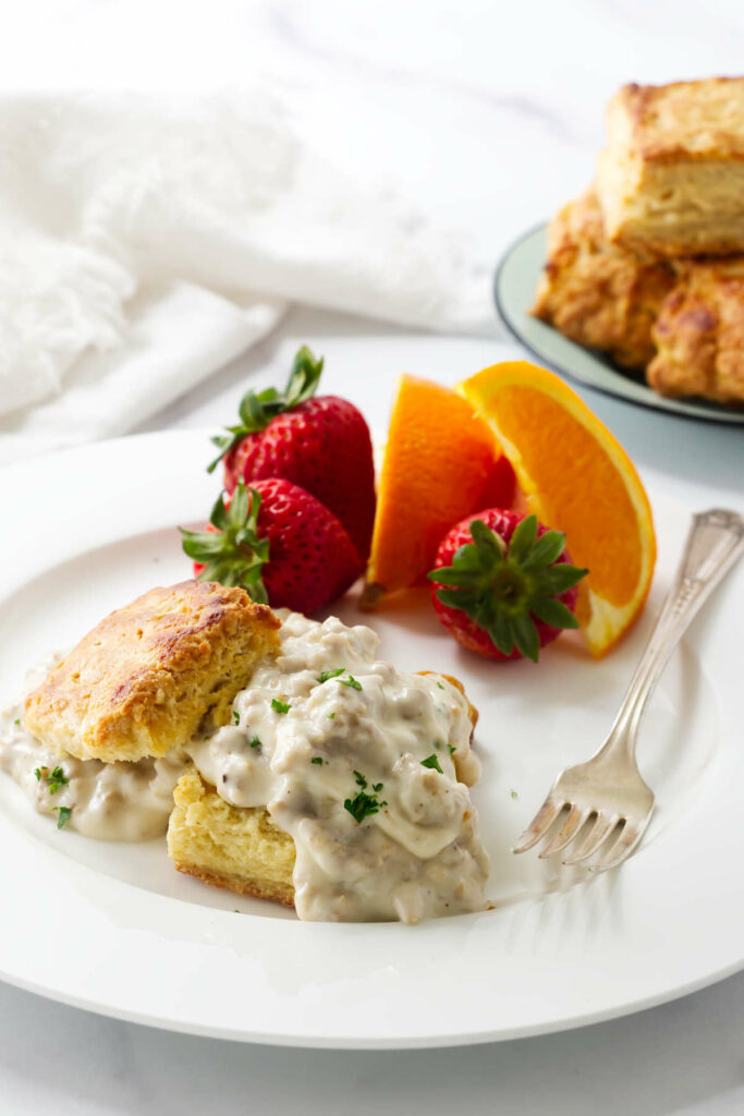 Sausage gravy and biscuits on a white plate with a stack of biscuits in the back.