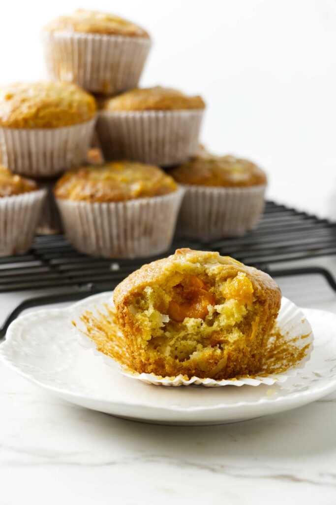 A partially eaten mango coconut muffin with a stack of muffins in the background.