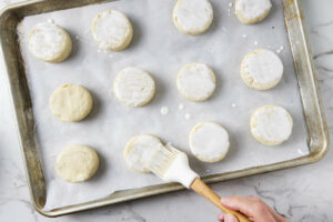 Brushing cream on top of the biscuits on a sheet pan.