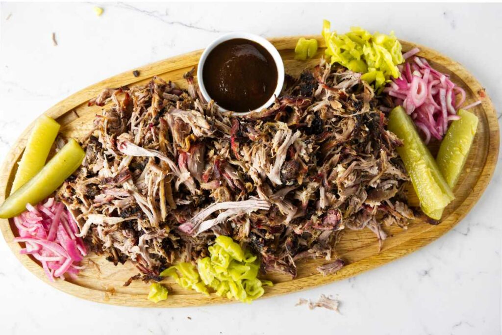 A cutting board with pulled pork butt and pickled veggies and bbq sauce.