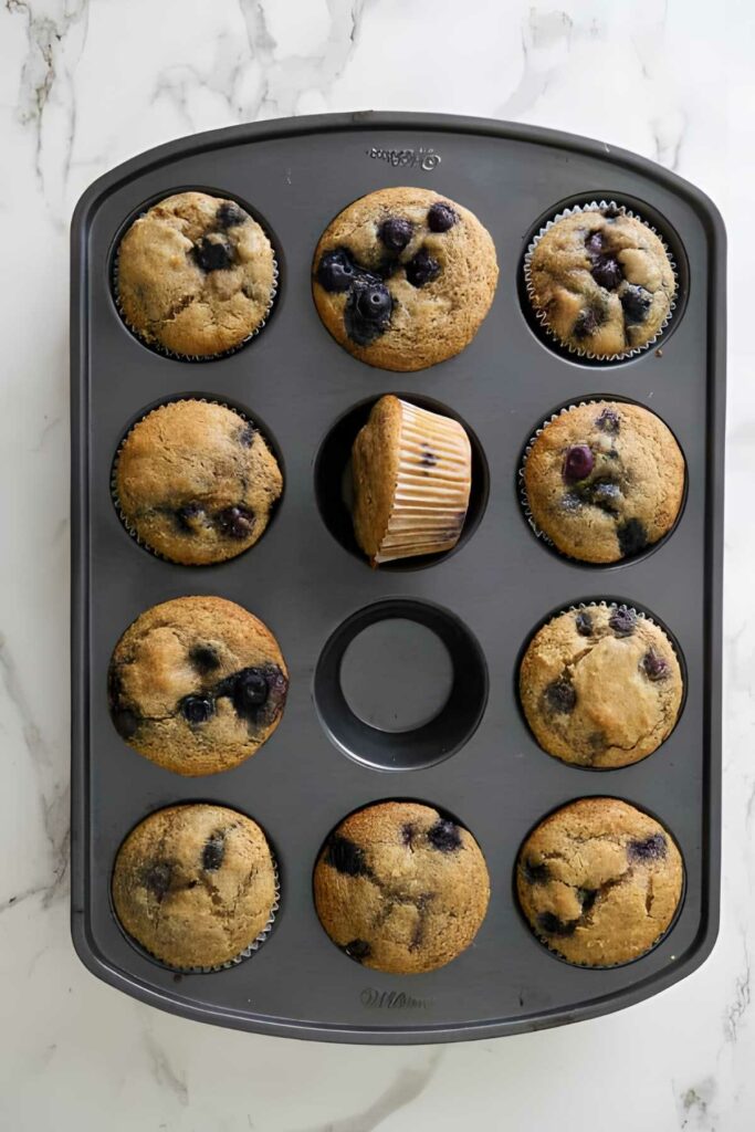 Muffin tray with 11 blueberry muffins, one tilted, and an empty slot.