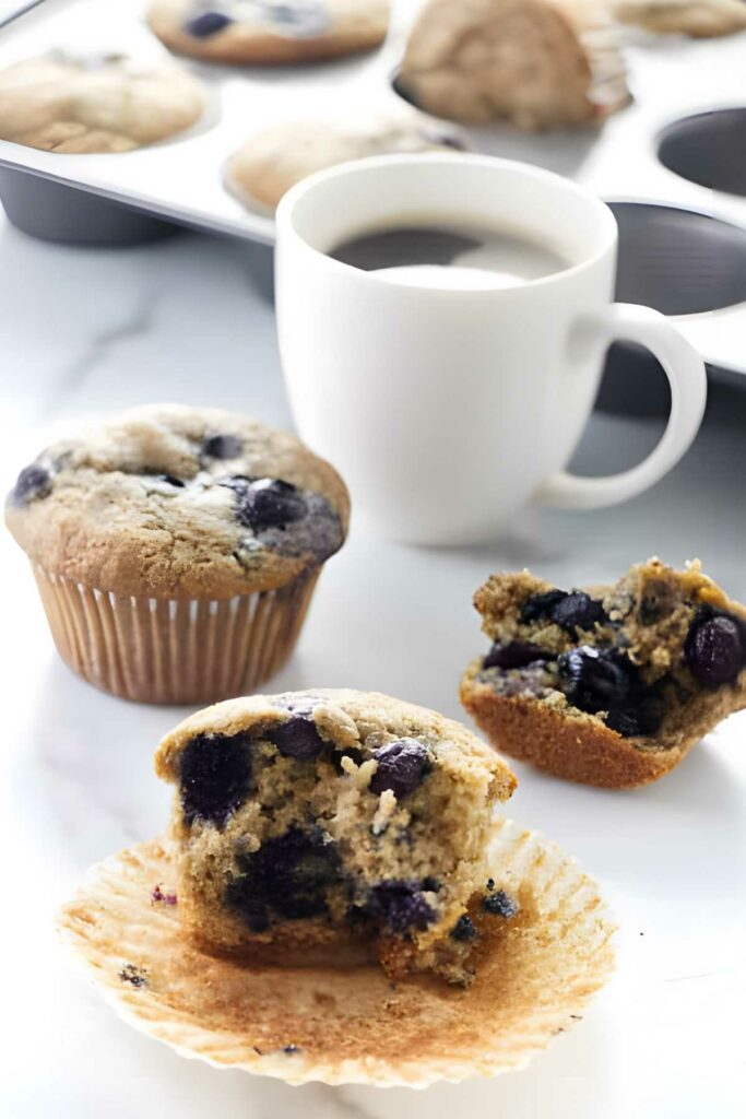 Blueberry muffin, halved muffin, coffee cup, and muffin tin on white surface.