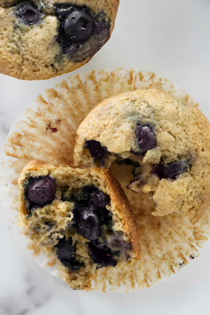 Broken spelt muffin on white liner, juicy blueberries inside.